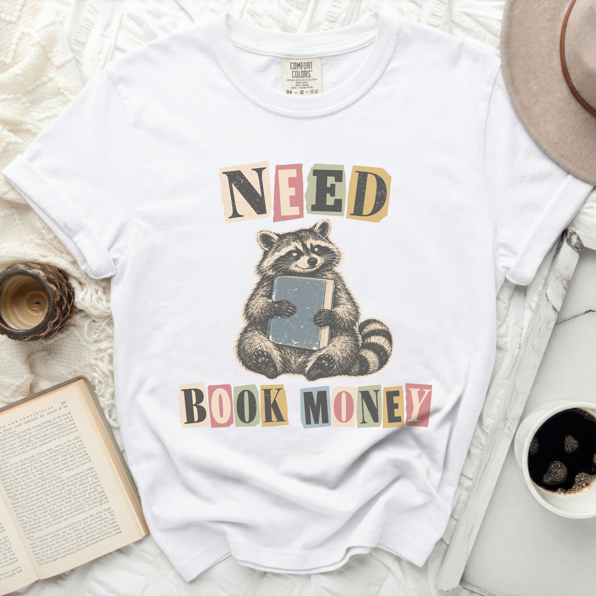 White t-shirt with raccoon holding book and text "NEED BOOK MONEY".