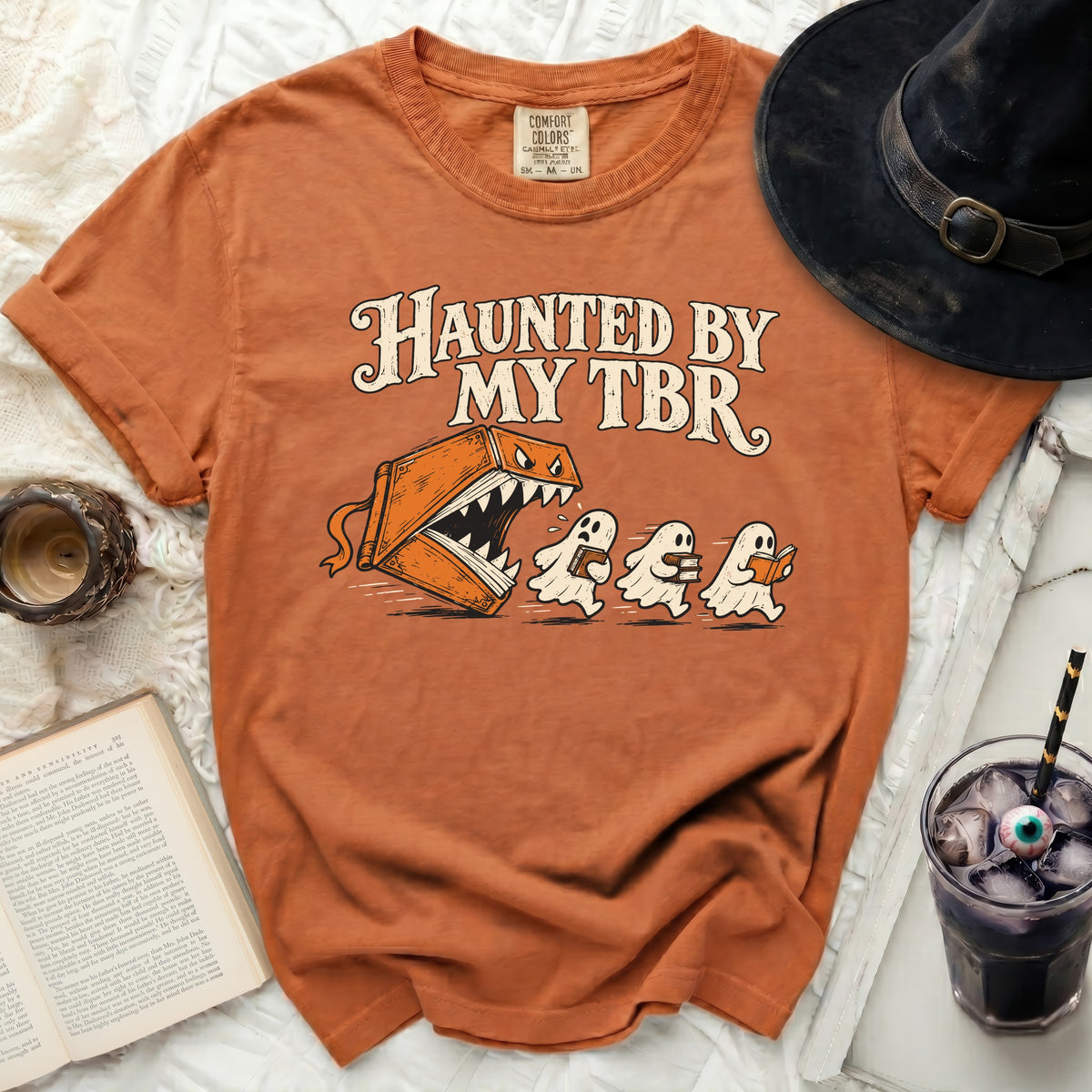 Orange t-shirt with "Haunted By My TBR" text and a book monster chasing ghosts.