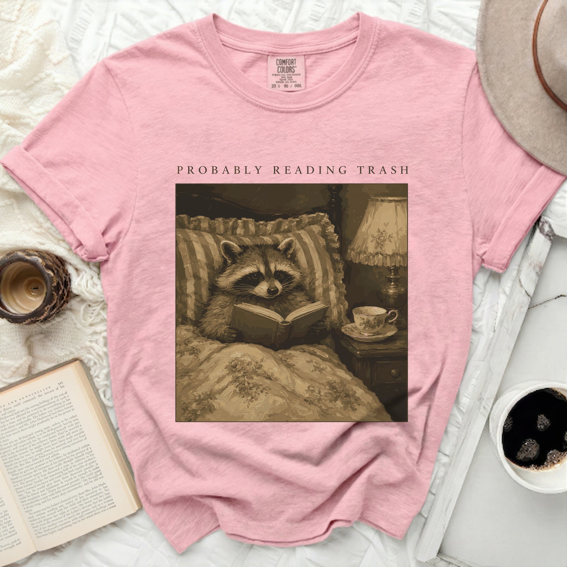 Pink t-shirt with illustration of raccoon reading in bed, text "PROBABLY READING TRASH".