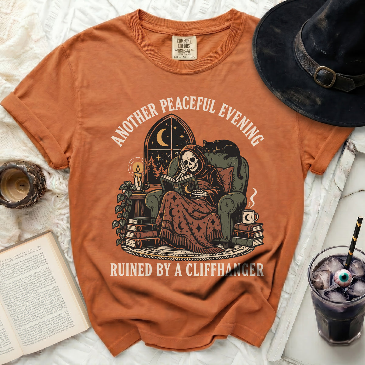 Orange t-shirt with a skeleton reading a book, text reads "Another peaceful evening ruined by a cliffhanger".