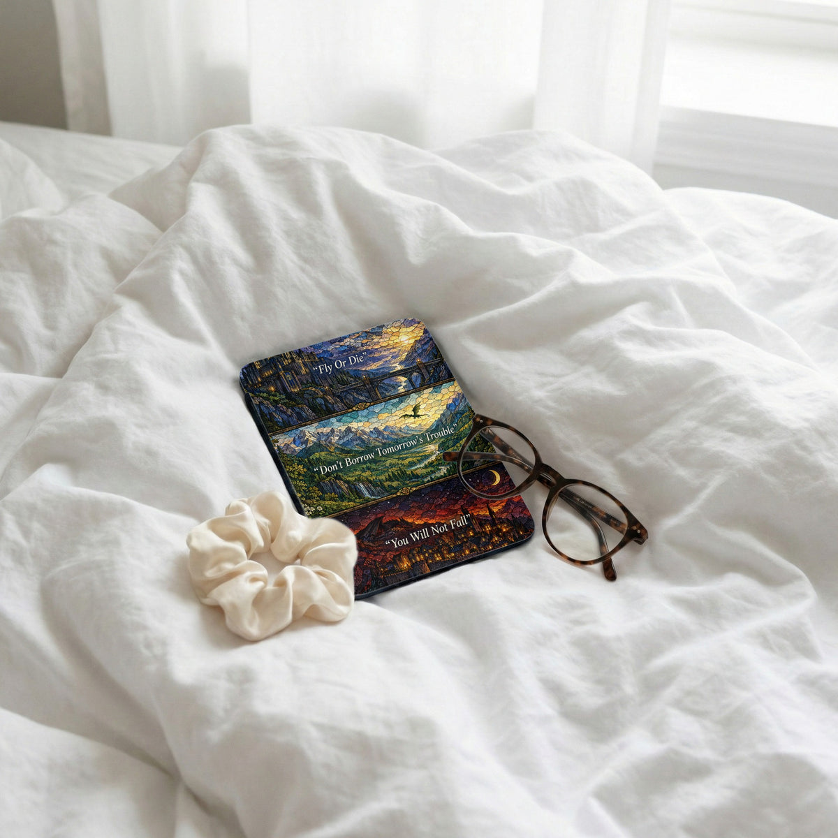 A Kindle case featuring stained-glass fantasy artwork rests on a white comforter beside glasses and a scrunchie.