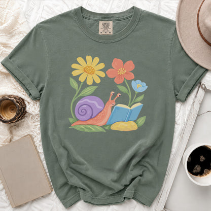 Green t-shirt with a watercolor print of a snail reading a book among flowers.