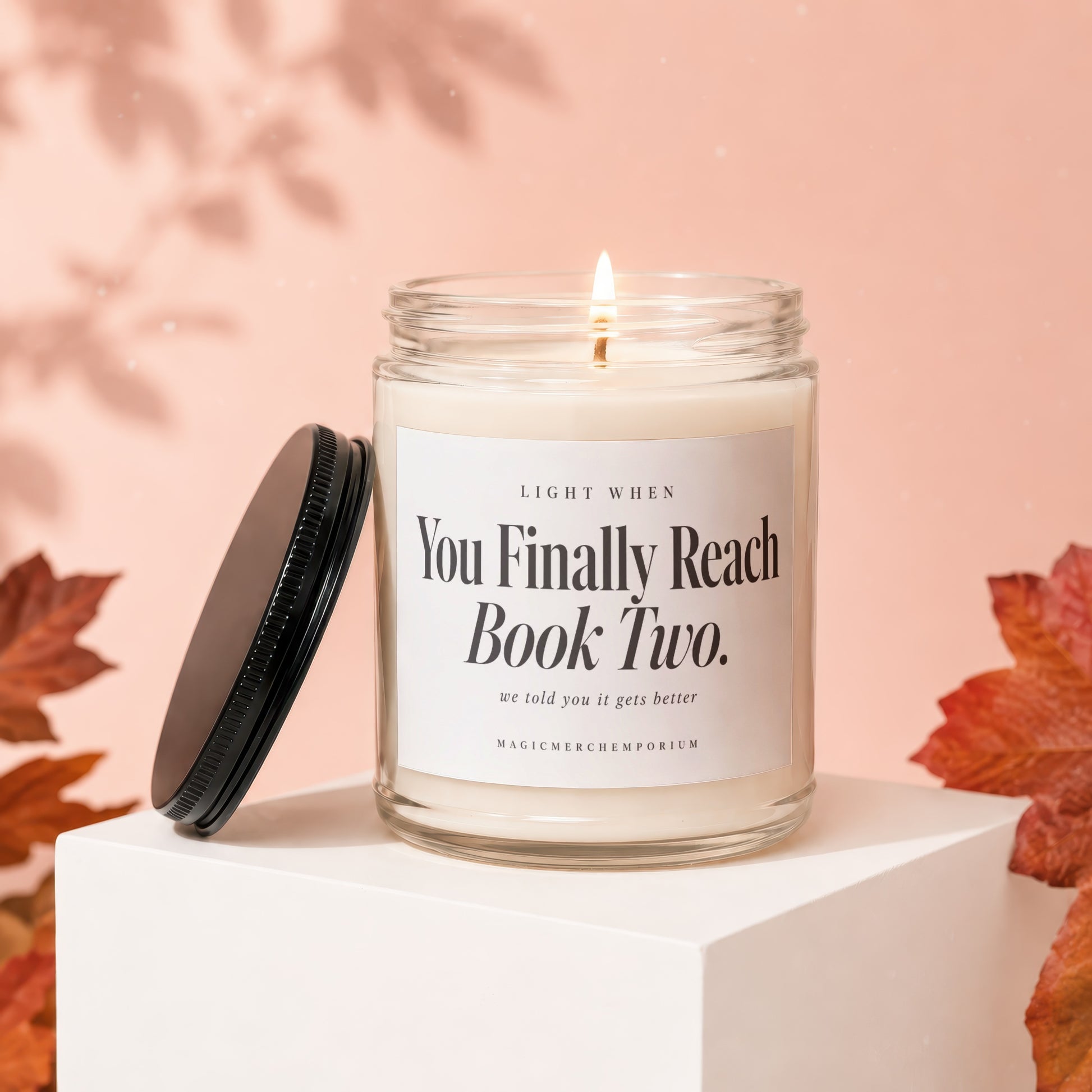 A lit candle in a glass jar with a black lid and white label rests on a white block against a peach background with autumn leaves.