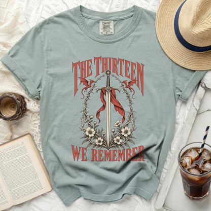 Light blue t-shirt with a sword, dragons, and flowers graphic reading "The Thirteen We Remember".