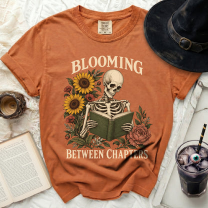 Orange t-shirt with skeleton reading a book and text "Blooming Between Chapters".