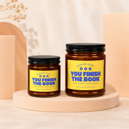 Two amber glass candles with yellow labels reading "You Finish The Book" on display.