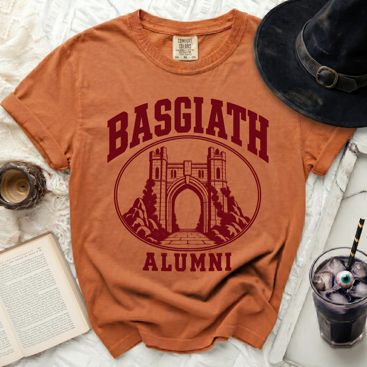 An orange crewneck t-shirt featuring a maroon graphic of a stone castle gate and the text Basgiath Alumni.