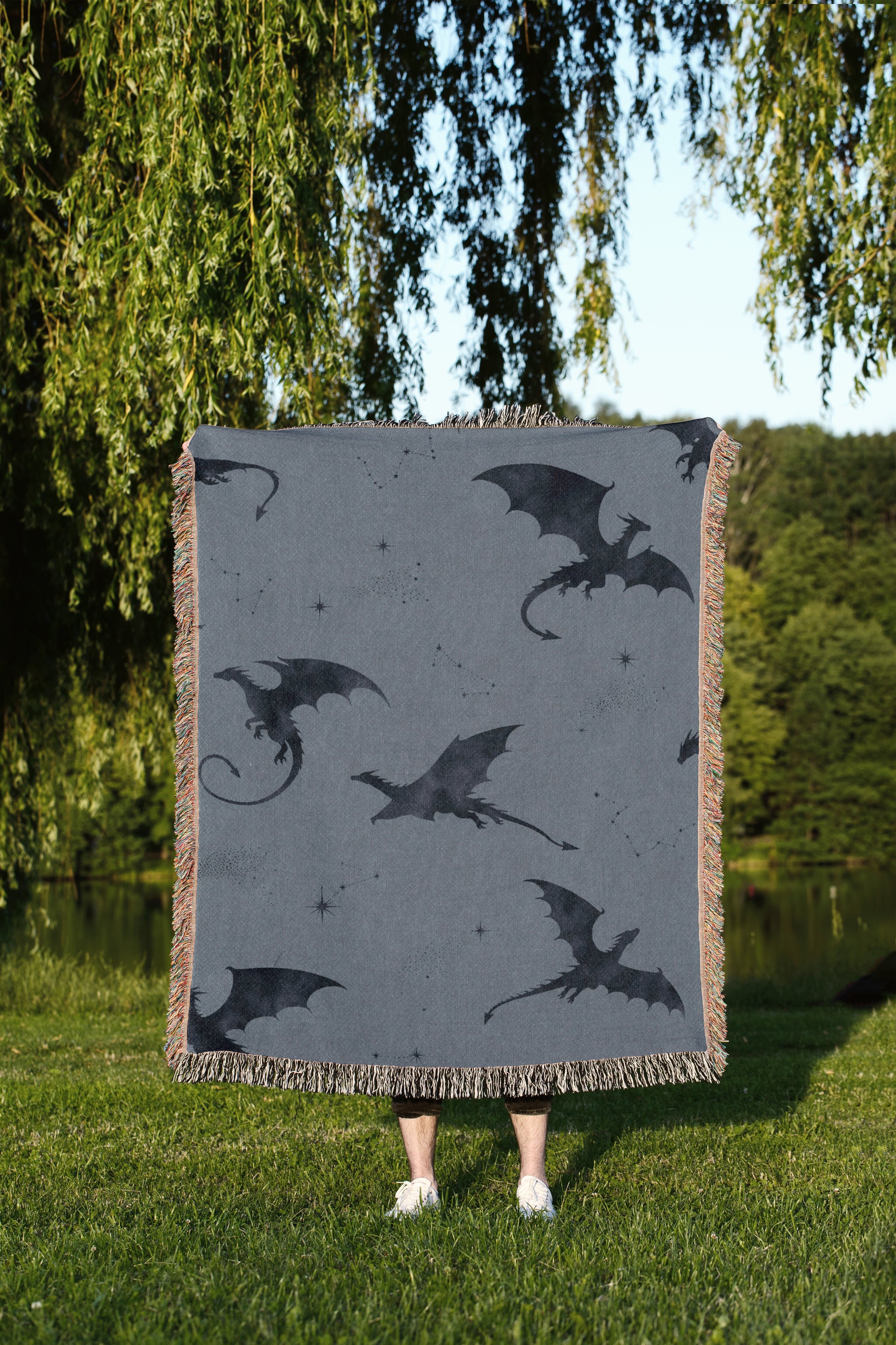A person is standing in a grassy area, holding a large square blanket with a dragon design on it.