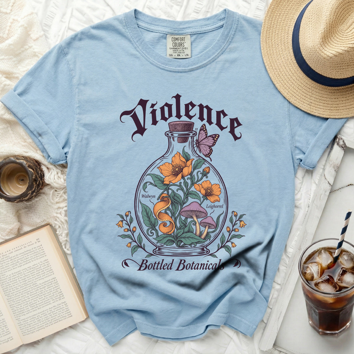 Light blue crewneck t-shirt featuring a graphic of flowers and mushrooms inside a glass bottle, text reading Violence Bottled Botanicals, surrounded by a straw hat, book, drink, and candle.
