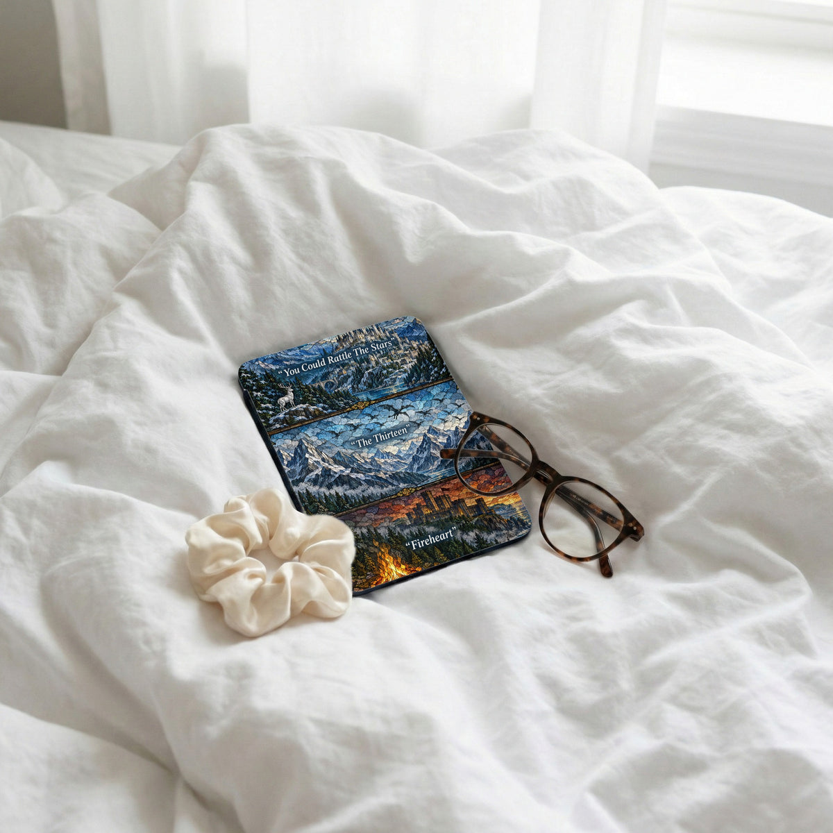 A Kindle case with stained glass artwork rests on white rumpled bedsheets beside tortoiseshell glasses and a white scrunchie.