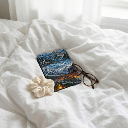 A Kindle case with stained glass artwork rests on white rumpled bedsheets beside tortoiseshell glasses and a white scrunchie.