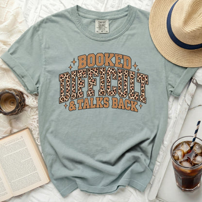 Dusty blue t-shirt with "Booked Difficult & Talks Back" leopard print graphic.