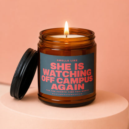 Amber jar candle with lit flame, lid off, and text "SHE IS WATCHING OFF CAMPUS AGAIN".