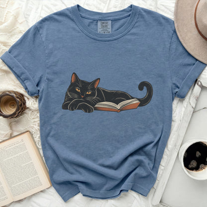 Black cat lying on an open book with Comfort Colors tag visible on shirt.
