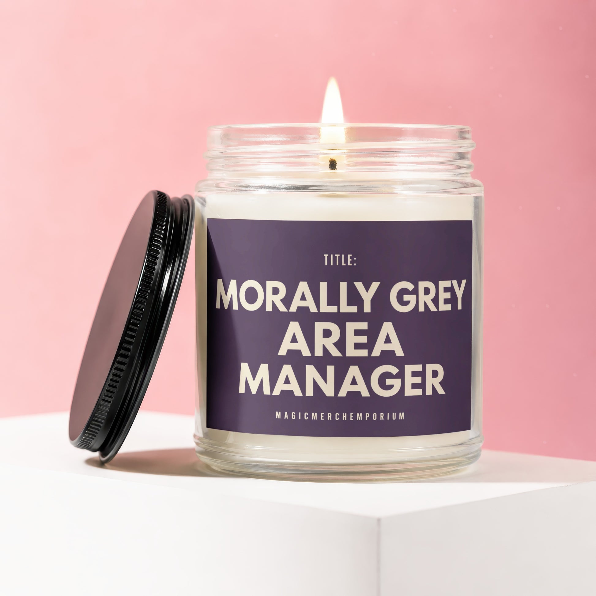 Lit candle in glass jar with lid off, reads "Morally Grey Area Manager"