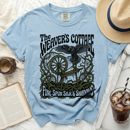Light blue t-shirt with "The Weaver's Cottage" crow and spinning wheel design.