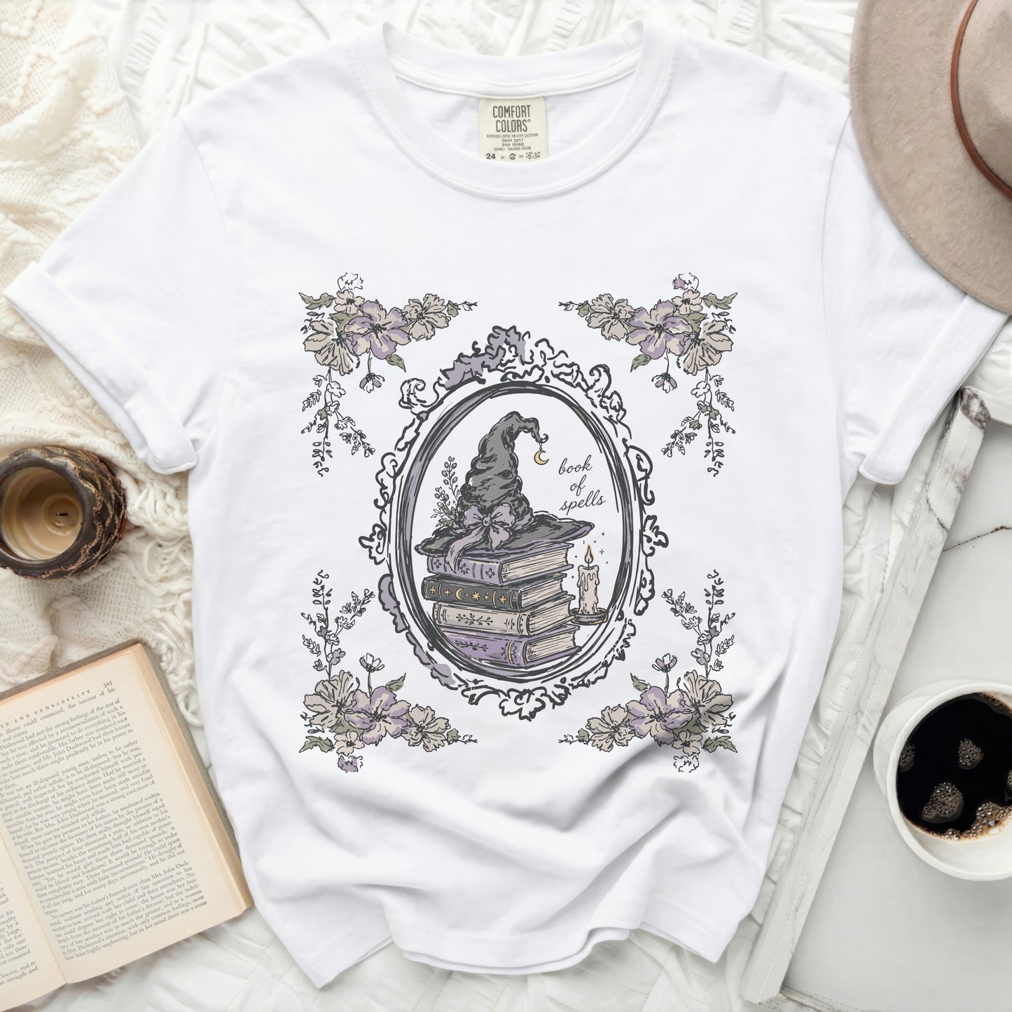 White t-shirt with a witch hat, stack of books, candle, and flowers graphic.