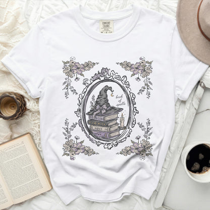 White t-shirt with a witch hat, stack of books, candle, and flowers graphic.
