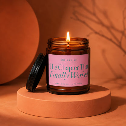 Lit amber glass candle with pink label reading "The Chapter That Finally Worked".