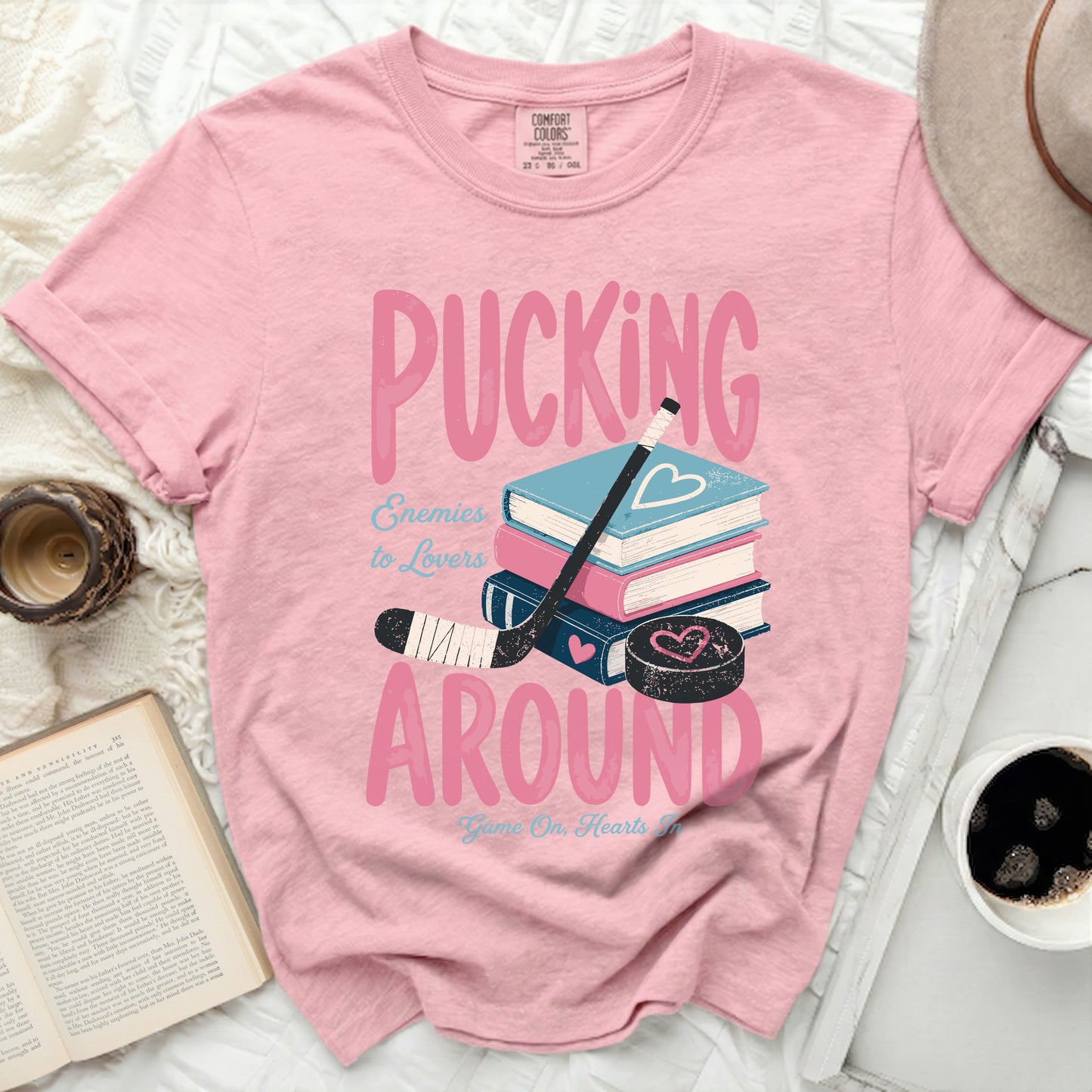 Pink t-shirt with "Pucking Around Enemies to Lovers" text and hockey stick.
