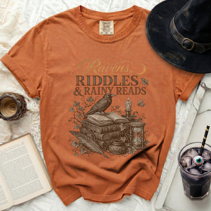 Orange comfort colors t-shirt with "Ravens, Riddles & Rainy Reads" graphic.