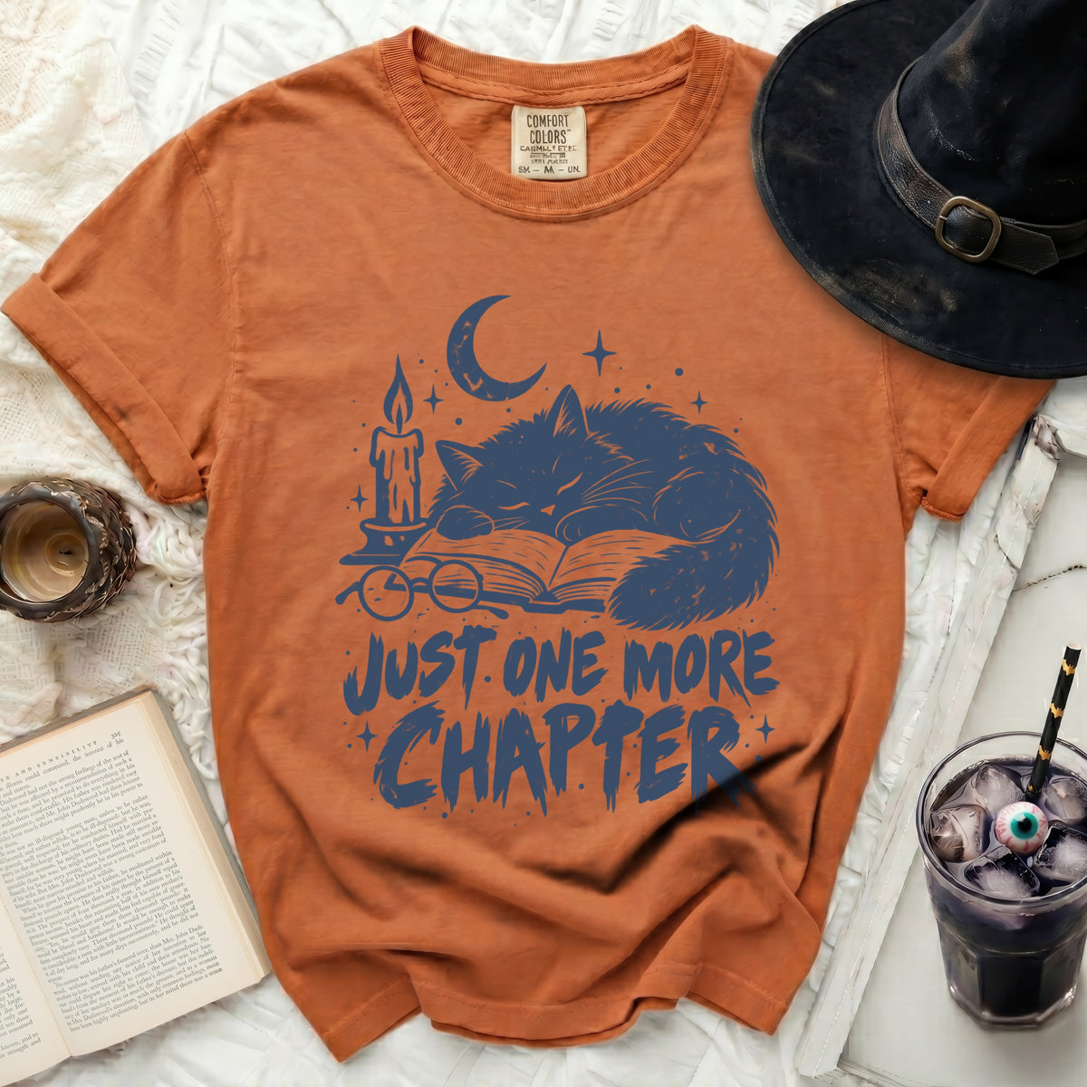 Orange t-shirt with blue graphic of a sleeping cat, book, candle, and text "JUST ONE MORE CHAPTER".