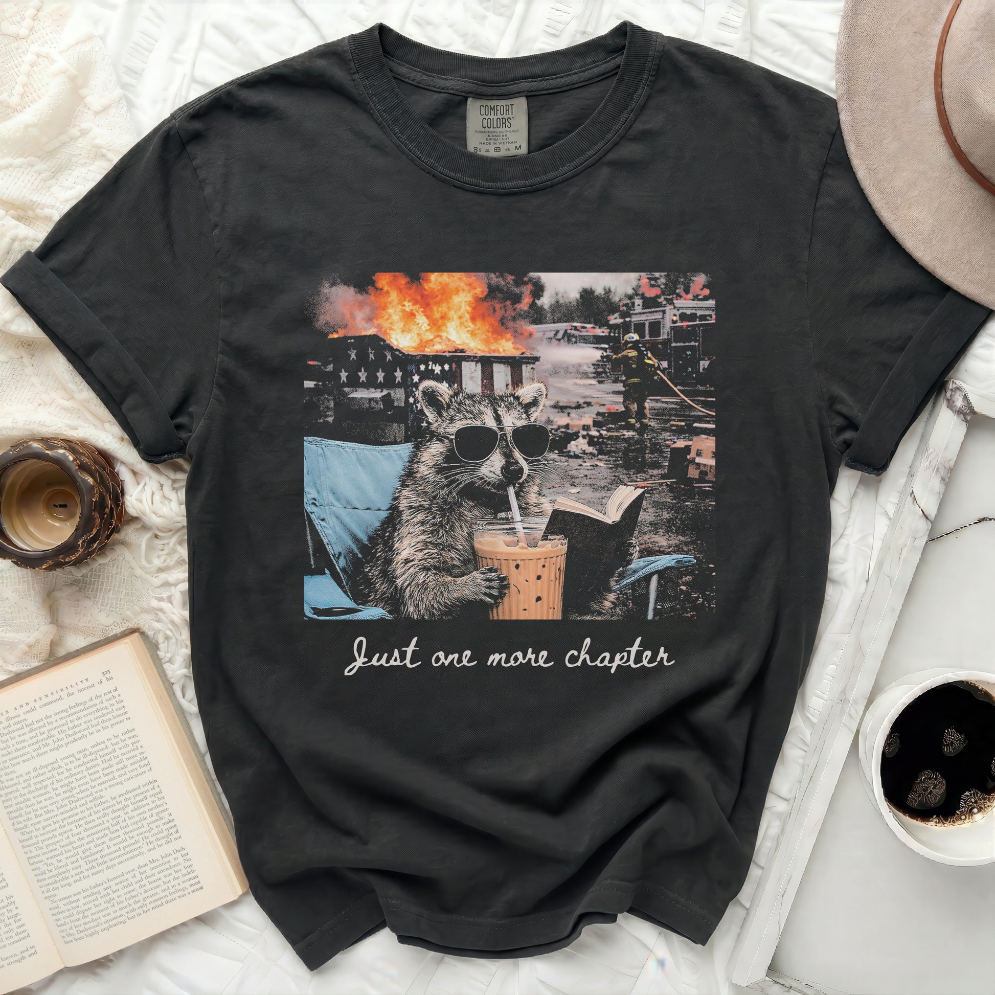 T-shirt with raccoon in a chair, coffee, and fire, reads "Just one more chapter".