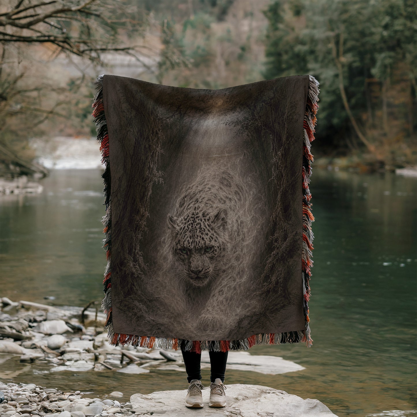 A person stands on a rock, holding a large blanket with a picture of a leopard on it, in front of a body of water surrounded by trees.
