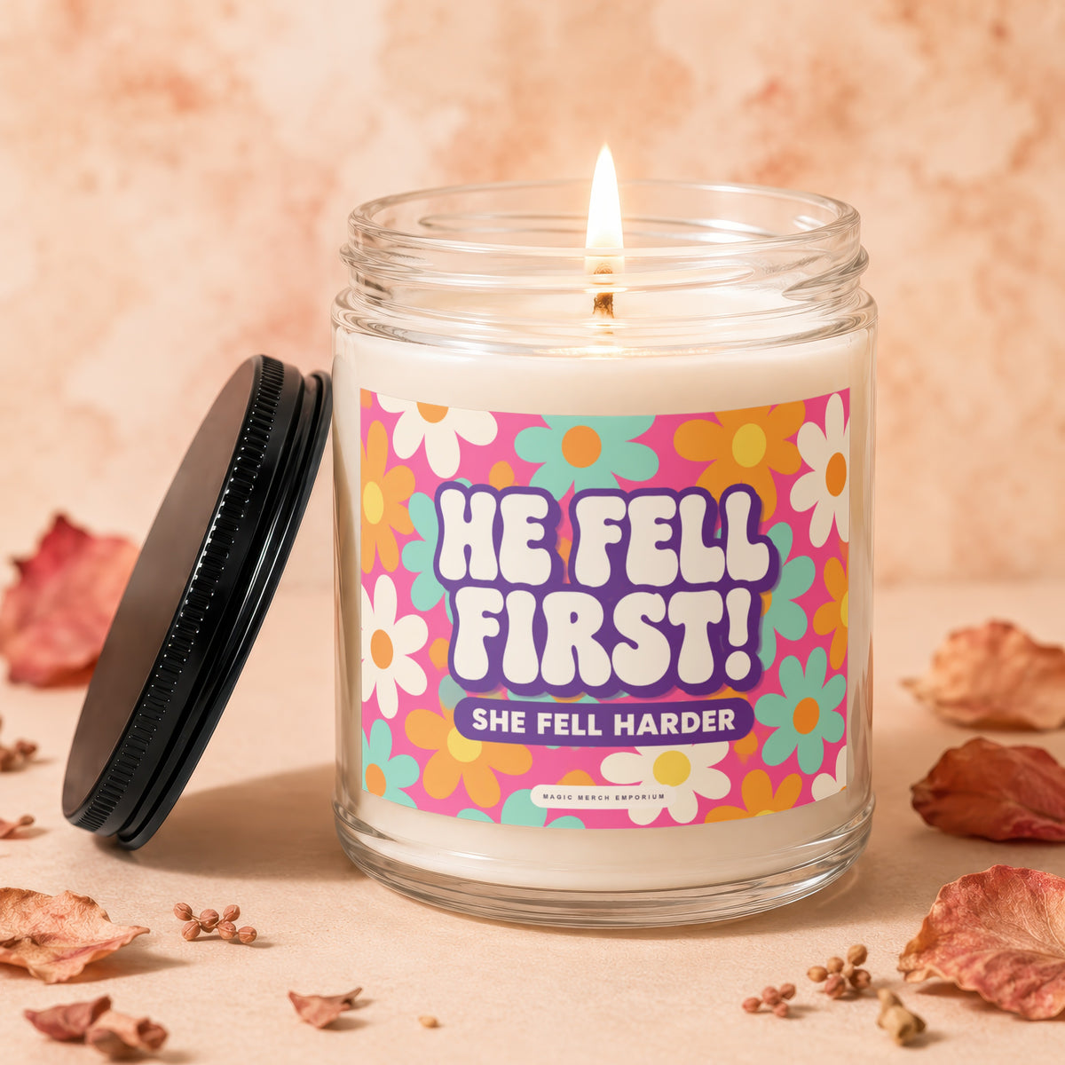 Lit candle with a label reading "HE FELL FIRST! SHE FELL HARDER" surrounded by dried flowers.