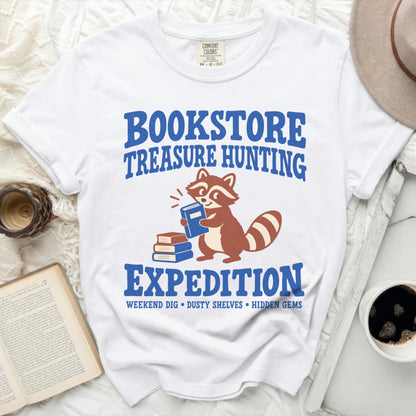 White t-shirt with blue text "BOOKSTORE TREASURE HUNTING EXPEDITION" and a raccoon holding a book.