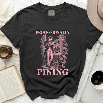 Dark t-shirt with pink graphic of a man reading by a window, "Professionally Pining".