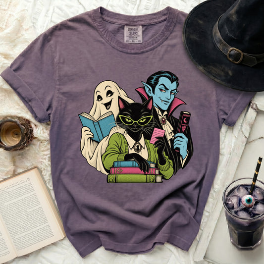 Purple t-shirt with graphic of a ghost, black cat, and vampire reading books.