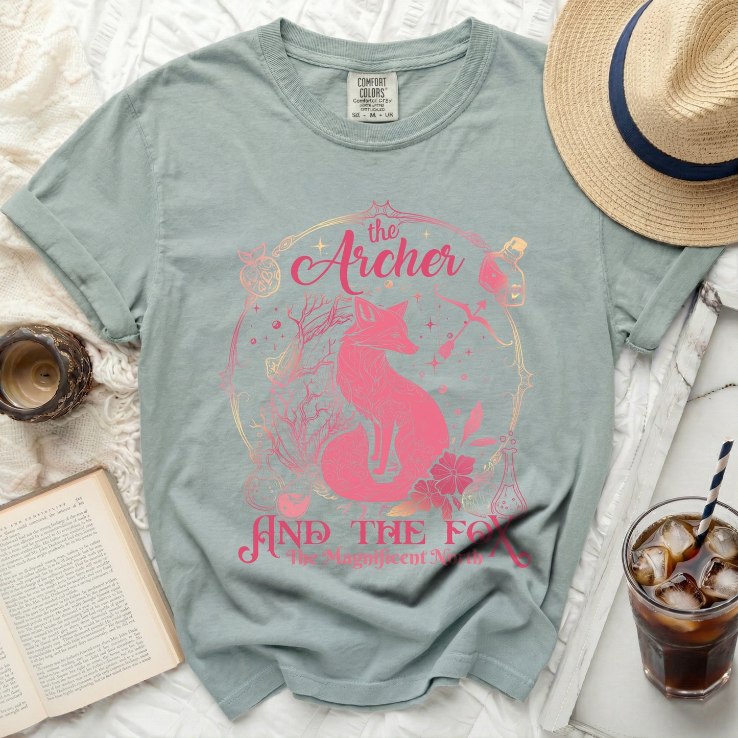 Pink fox and text graphic on a sage green t-shirt with a hat and book.