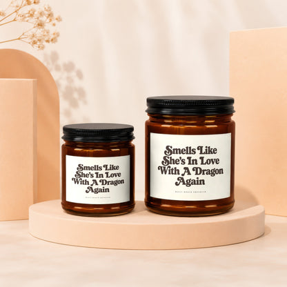 Two amber glass candles with black lids displaying the text "Smells Like She's In Love With A Dragon Again".
