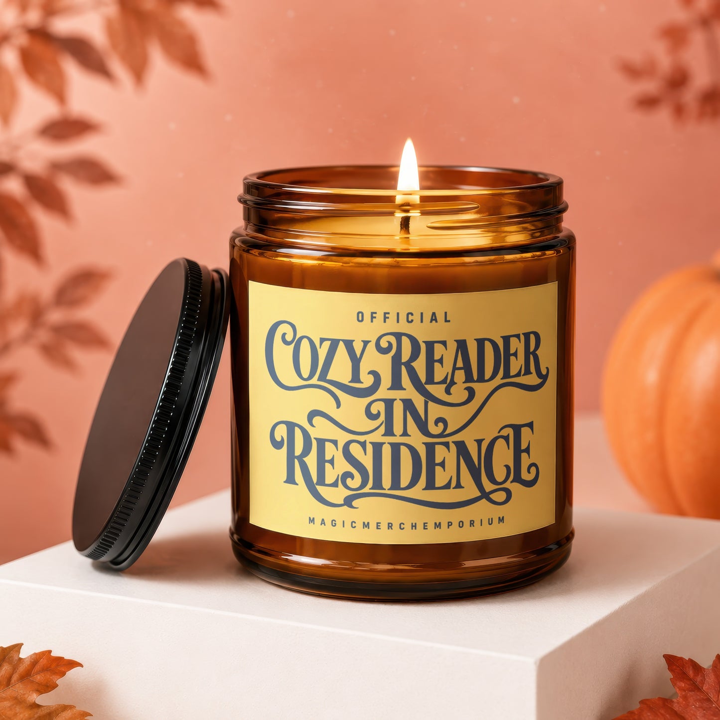 Amber glass candle with "Cozy Reader In Residence" label, lit wick, and pumpkin decor.