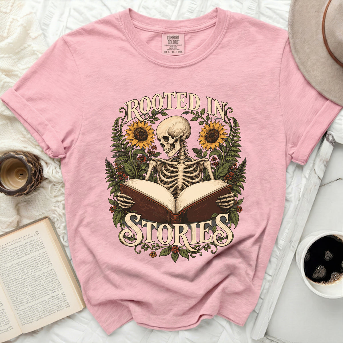 Pink t-shirt with a skeleton reading a book, surrounded by sunflowers and ferns, and the words "Rooted in Stories".
