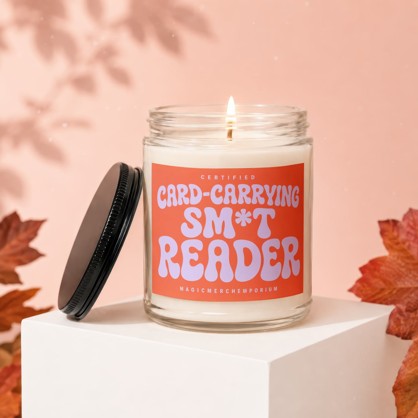 Candle with "Certified Card-Carrying Sm*t Reader" text on orange label. Lid is off.
