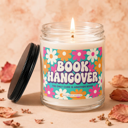 Glass candle with lit wick, labeled "Book Hangover," surrounded by dried leaves.