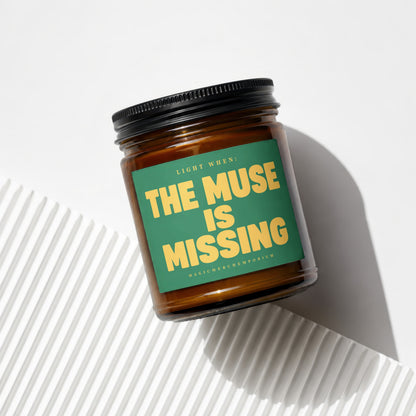 Amber jar candle with green label reading "THE MUSE IS MISSING"