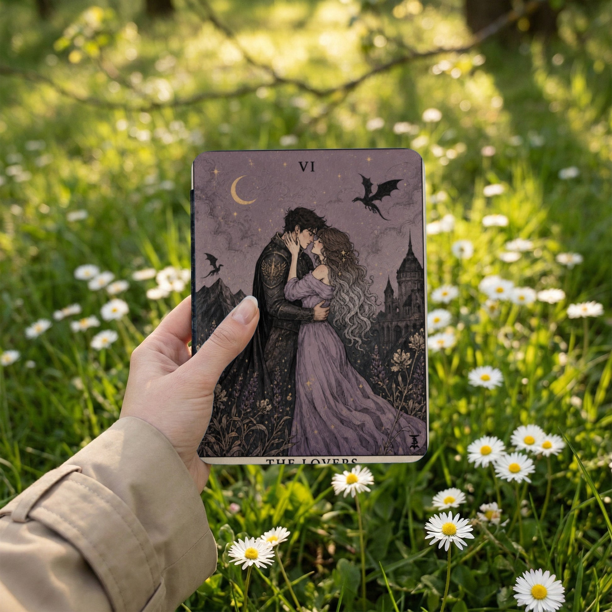 Tarot card "The Lovers" with a couple embracing, moon, and dragons, held in a hand outdoors.