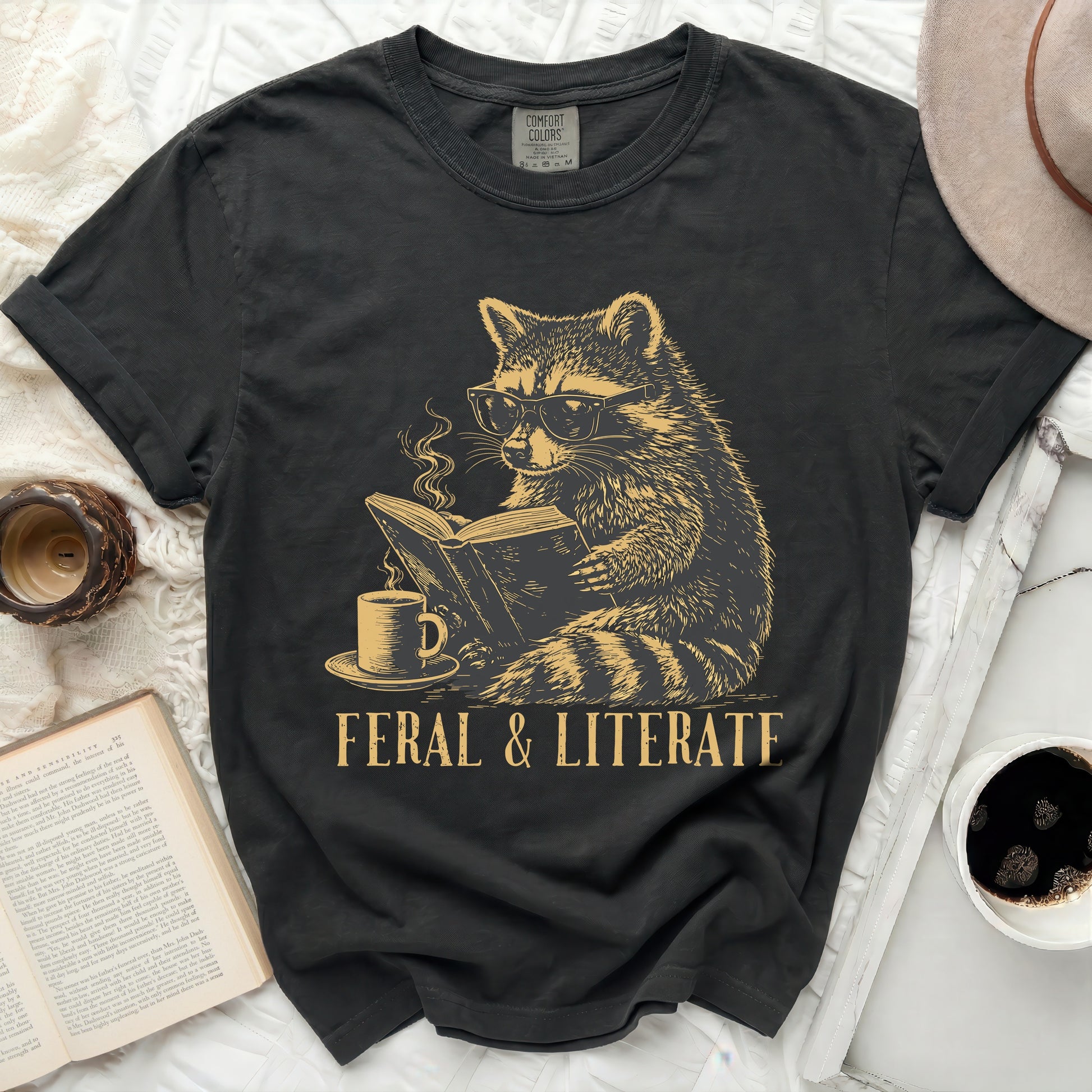 Dark t-shirt with gold graphic of a raccoon reading a book and drinking coffee.