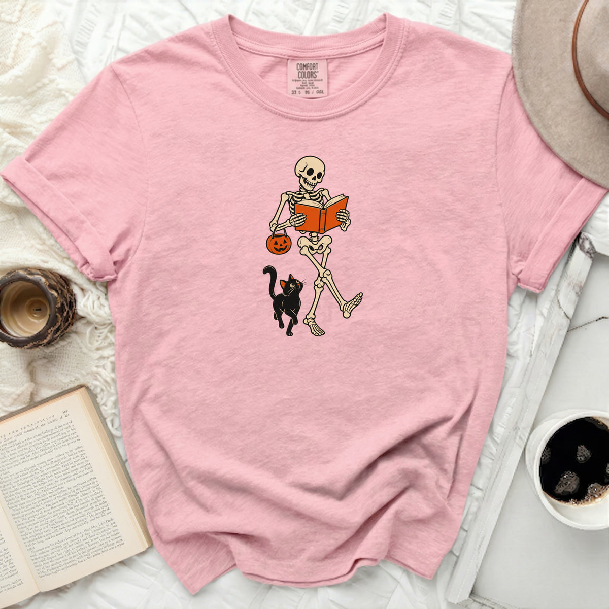 Pink t-shirt with graphic of skeleton reading book, holding pumpkin, with black cat.