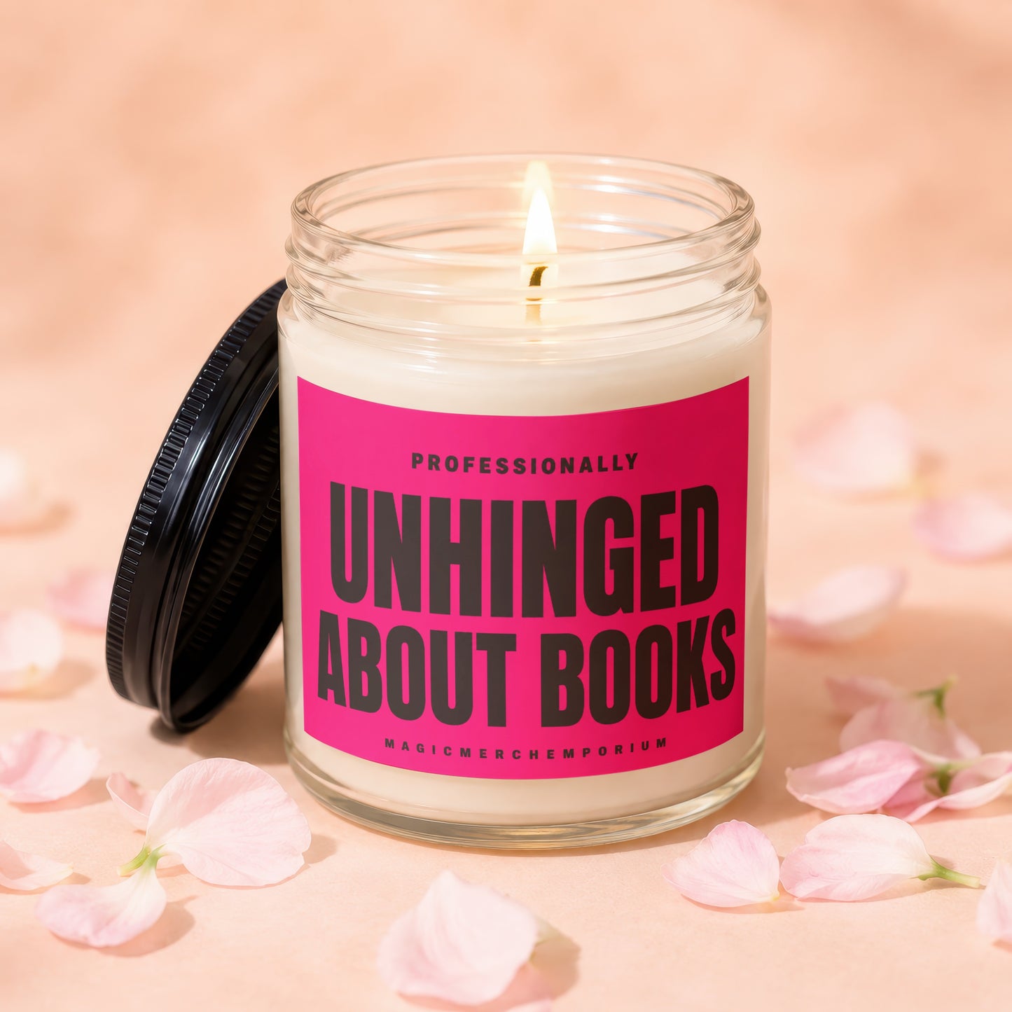 Glass candle with lit wick, black lid, and pink label reading "Professionally Unhinged About Books".