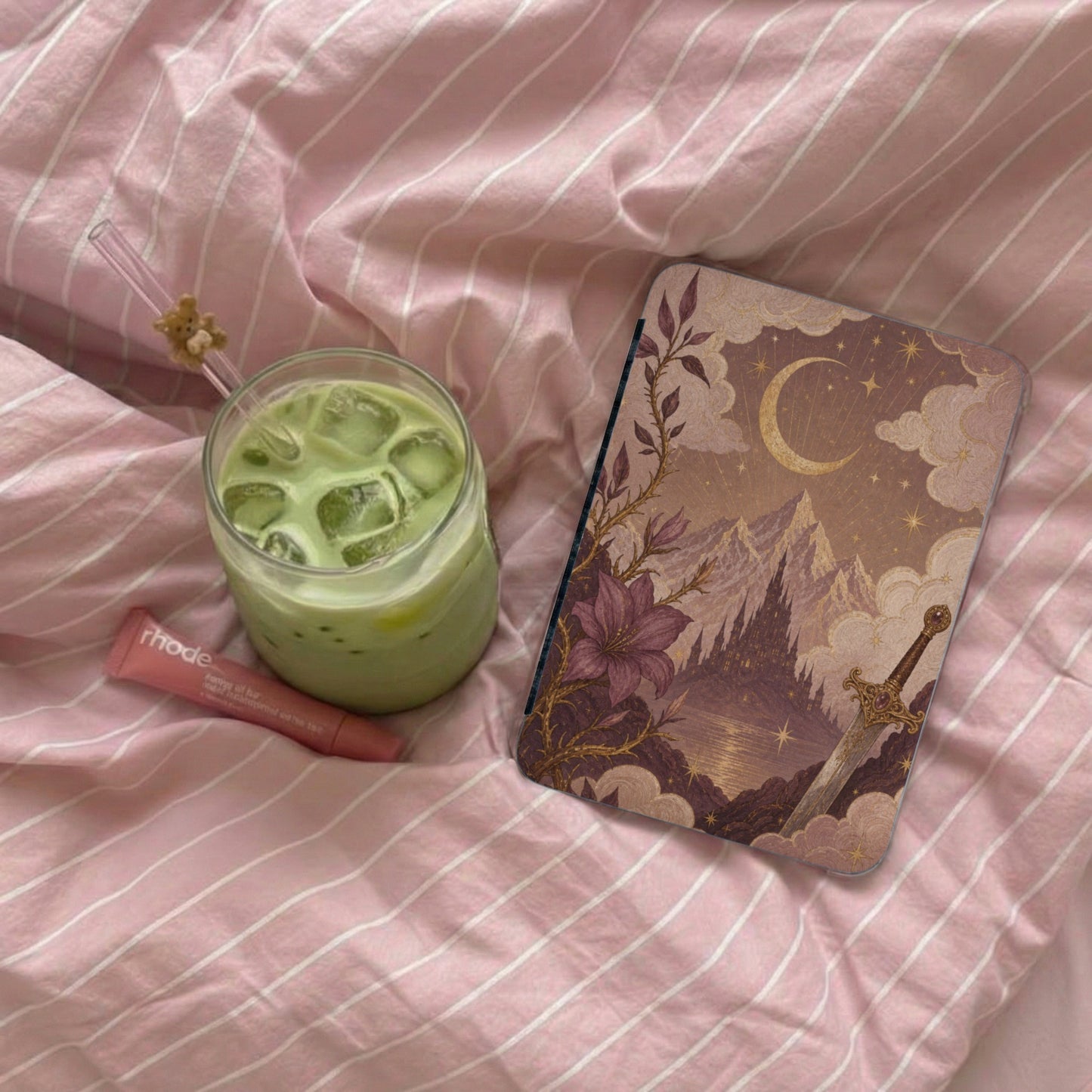 Ipad case with fantasy art, a glass of matcha, and lip balm on pink striped bedding.