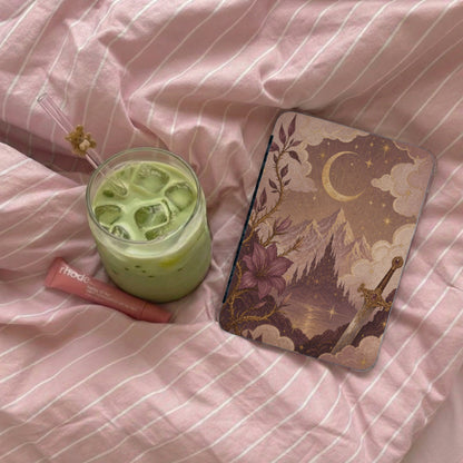 Ipad case with fantasy art, a glass of matcha, and lip balm on pink striped bedding.