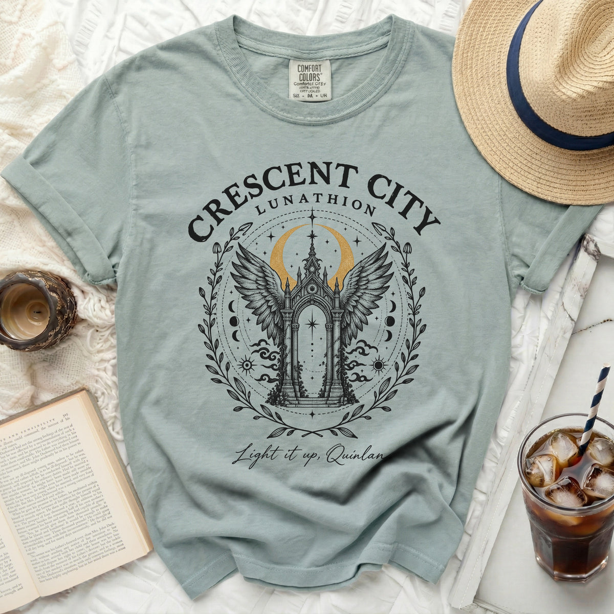 A sage green t-shirt features a black and gold Crescent City Lunathion graphic, surrounded by props.