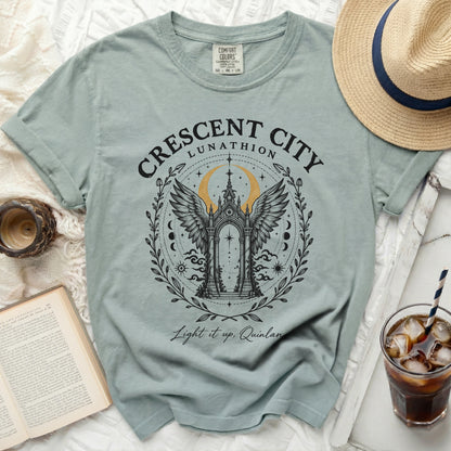 A sage green t-shirt features a black and gold Crescent City Lunathion graphic, surrounded by props.