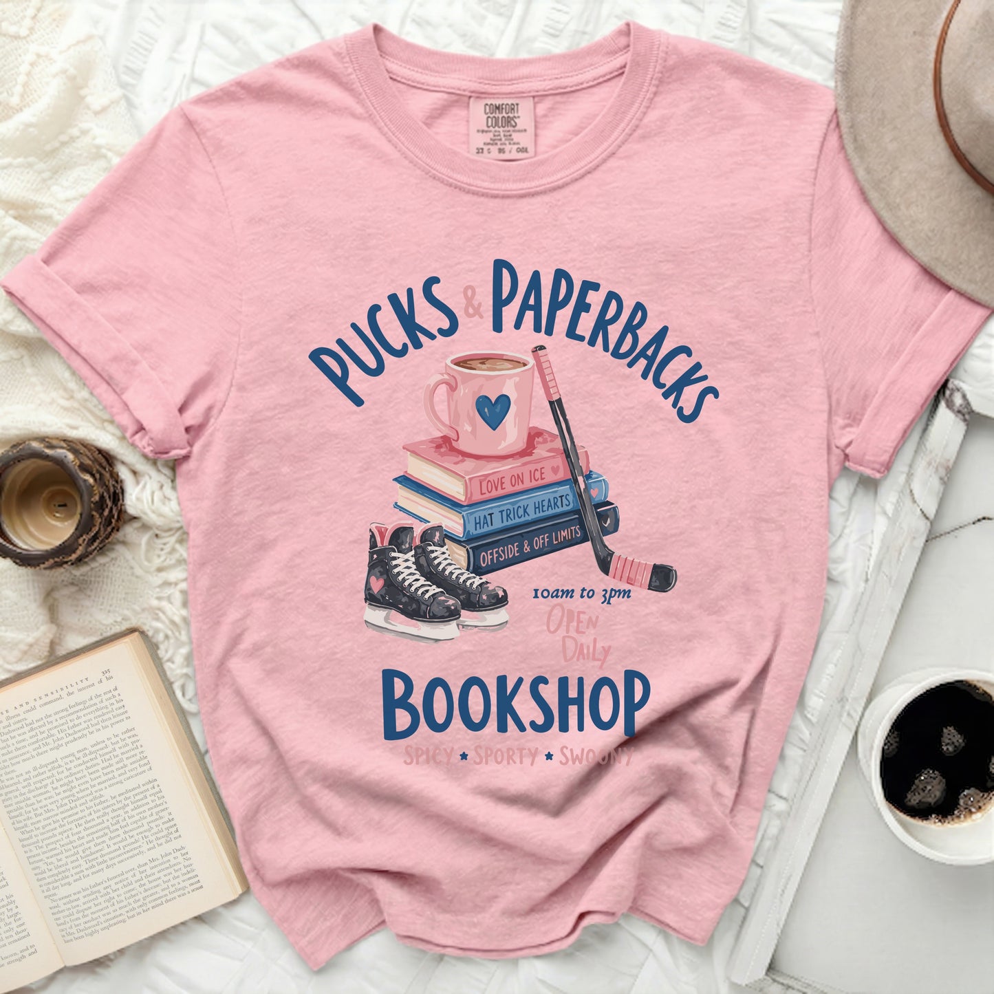 Pink t-shirt with "Pucks & Paperbacks" text, books, hockey skates, and stick.