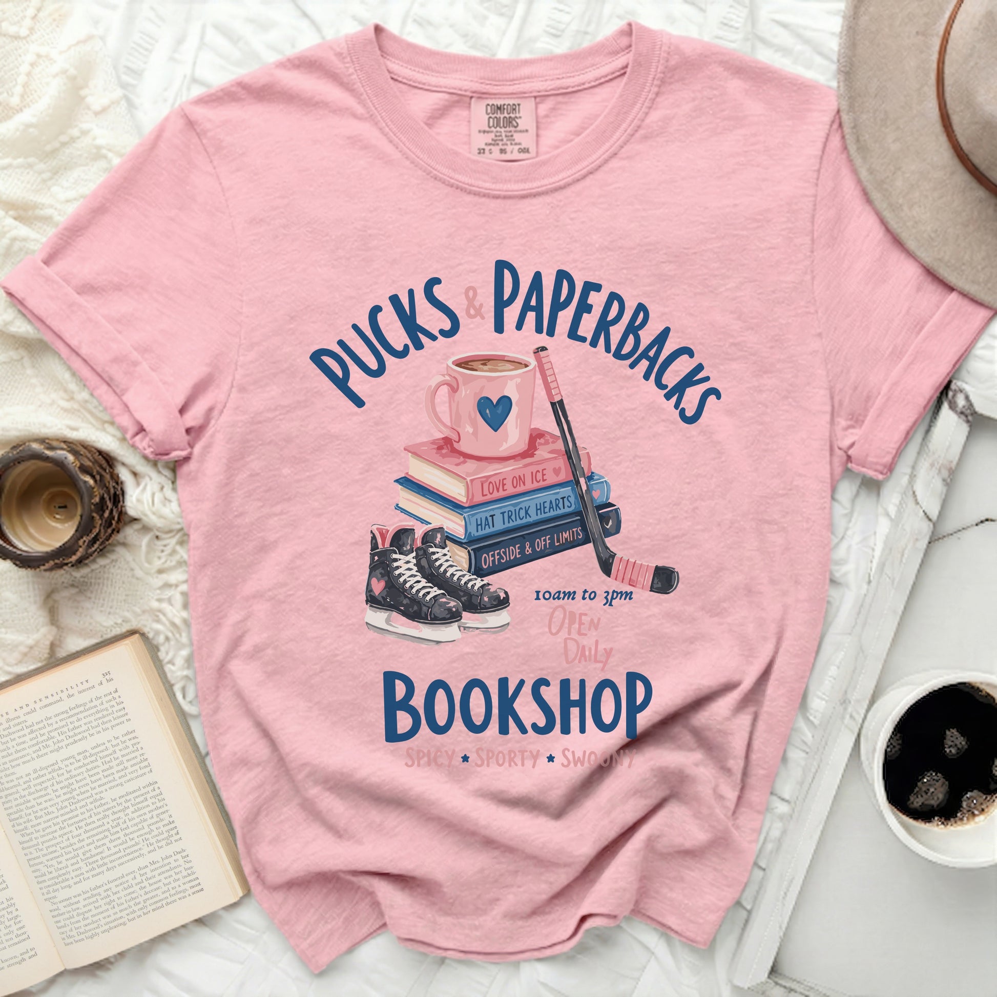 Pink t-shirt with "Pucks & Paperbacks" text, books, hockey skates, and stick.