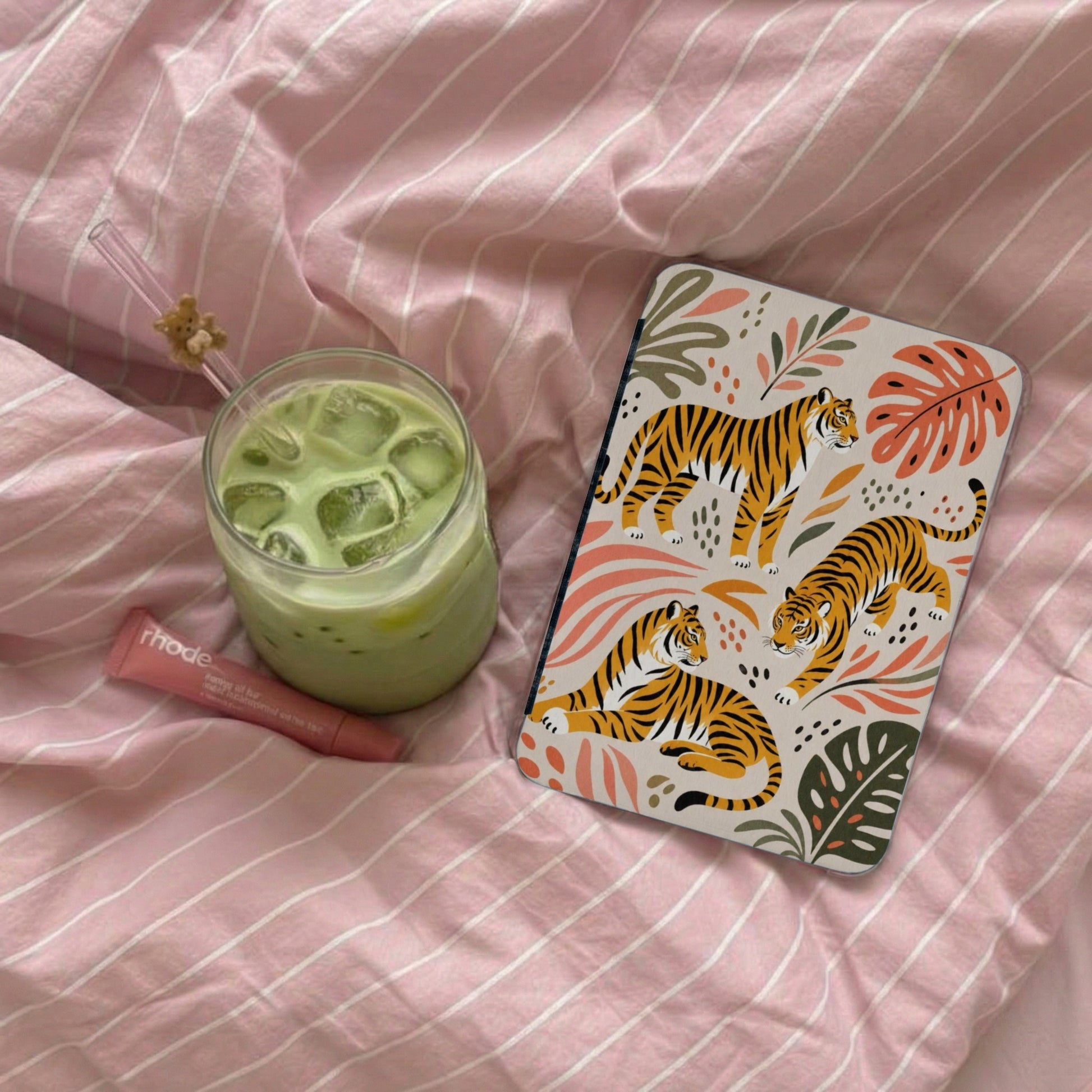 Journal with tiger print, iced green drink, and lip balm on pink striped bedding.